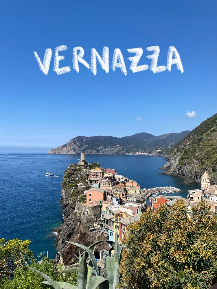 View of Vernazza, Cinque Terre. Orange and yellow-toned buildings flanked by the blue sea and green cliffs.