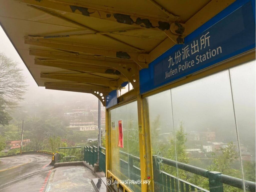 Yellow bus stop with nameplate Jioufen Police Station, surrounded by misty mountain